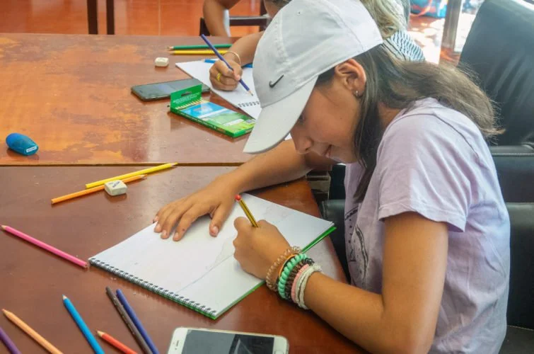 Una niña escribiendo en un cuaderno en una mesa de madera, rodeada de lápices, un teléfono móvil, y otros niños en el fondo, en un ambiente de aula.