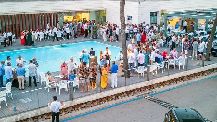 Evento social junto a una piscina en un área moderna con muchas personas, mesas y sillas, en un ambiente al aire libre.