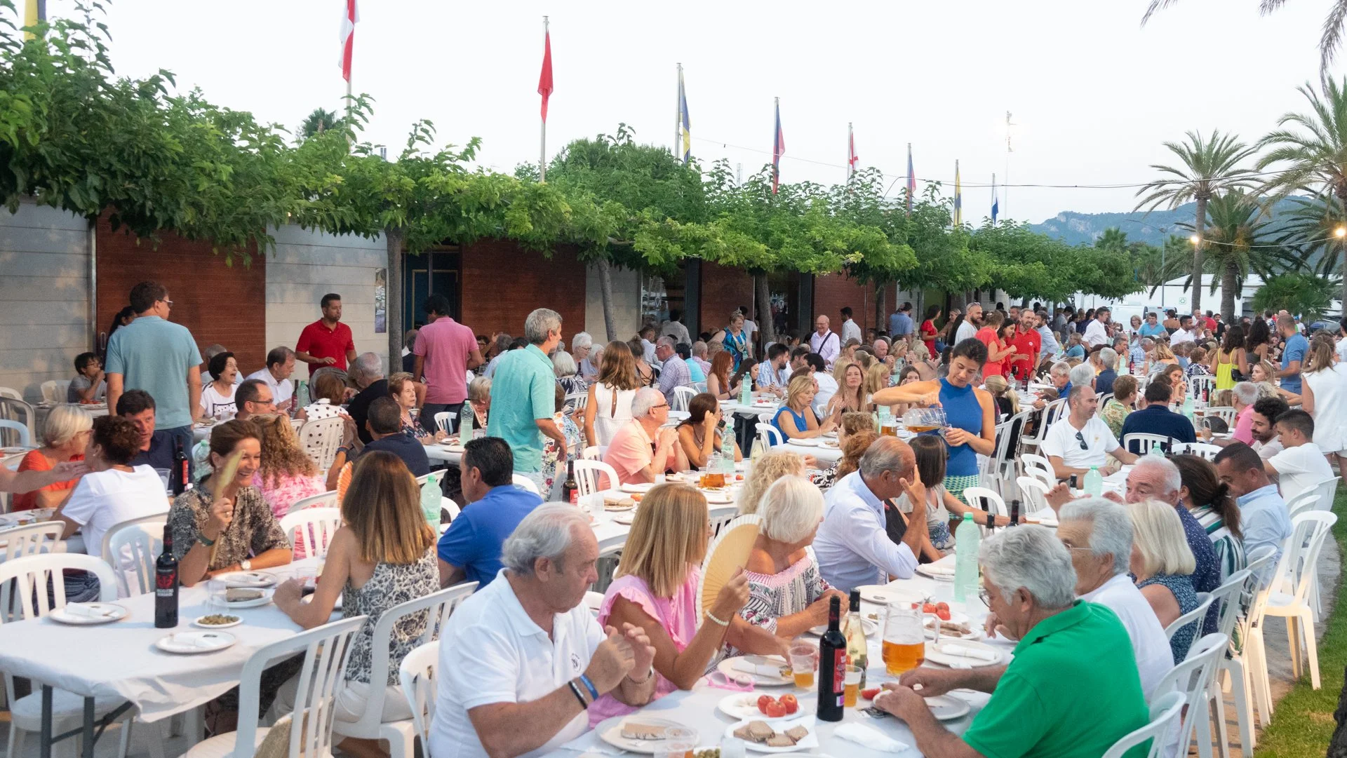 Una multitud de personas mayores y jóvenes en una fiesta al aire libre, disfrutando de comida y bebida en mesas con sillas blancas, con árboles y banderas en el fondo, y montañas al horizonte.