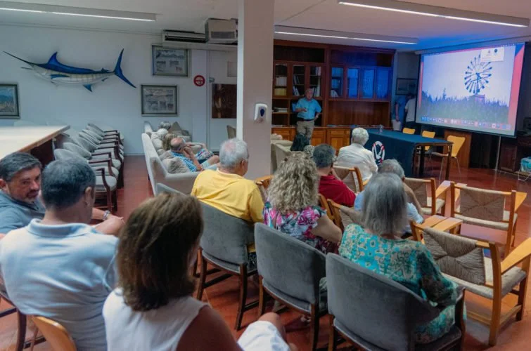 Personas sentadas viendo una presentación en una sala, en la pared hay un pez martillo decorativo y hay un proyector en la pantalla