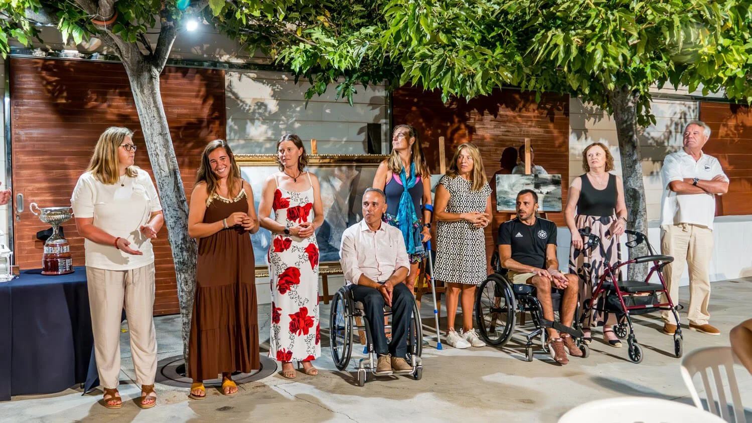 Grupo de personas con y sin discapacidad en un evento al aire libre bajo árboles con sombra, frente a un fondo de paredes de madera.