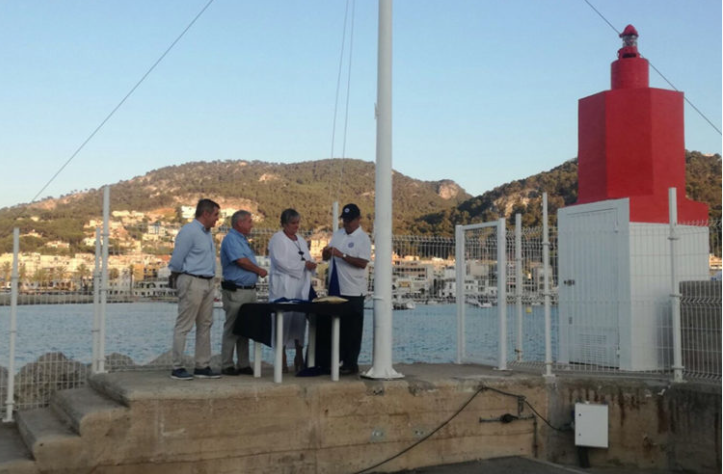 Cuatro personas en uniforme de chef con una mesa, cerca del mar y un faro rojo, en un muelle con fondo de montañas y casas.