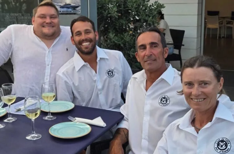 Cuatro personas sentadas en una mesa en un restaurante, sonriendo y vistiendo camisas blancas, algunas con logotipos de un club de yates.