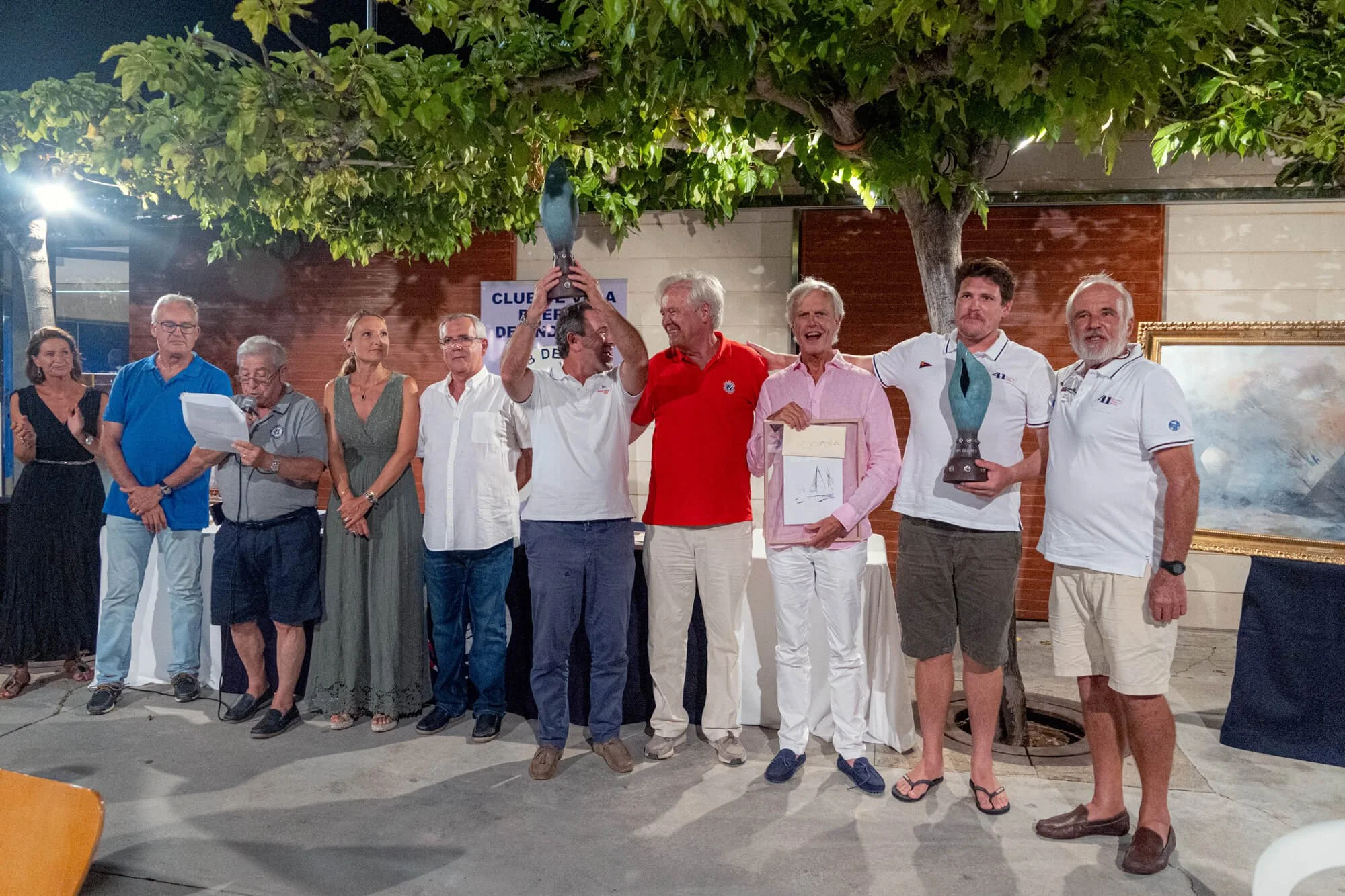 Grupo de personas en una ceremonia nocturna, algunas sosteniendo un trofeo y otros con reconocimientos, bajo un árbol iluminado.