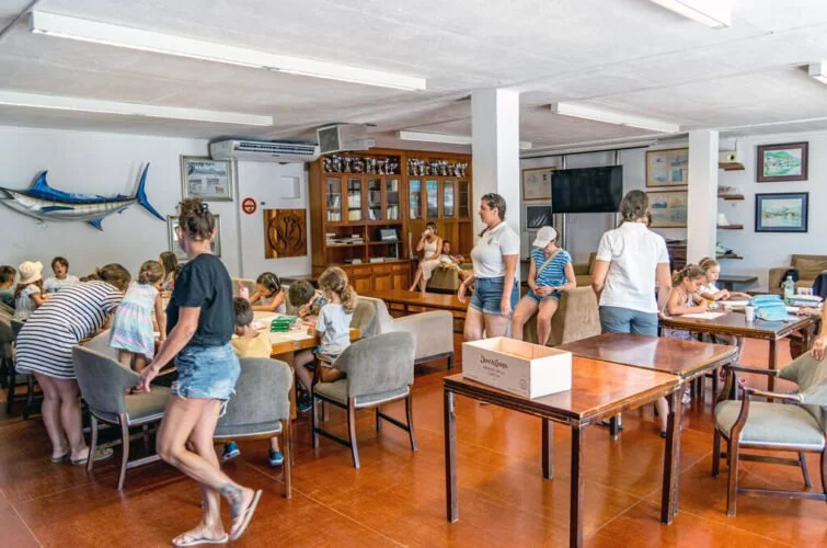 Niños y adultos en una sala con mesas y sillas, algunos niños dibujando, otros en actividades, paredes decoradas con pescado y cuadros, ambiente animado y organizado.