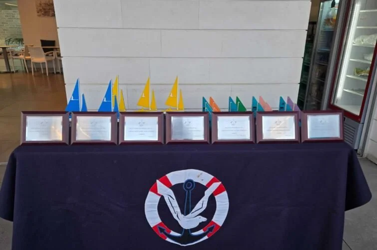 Mesa con premios y diplomas, decorada con un logotipo de ancla y una paloma, en un evento posiblemente náutico.
