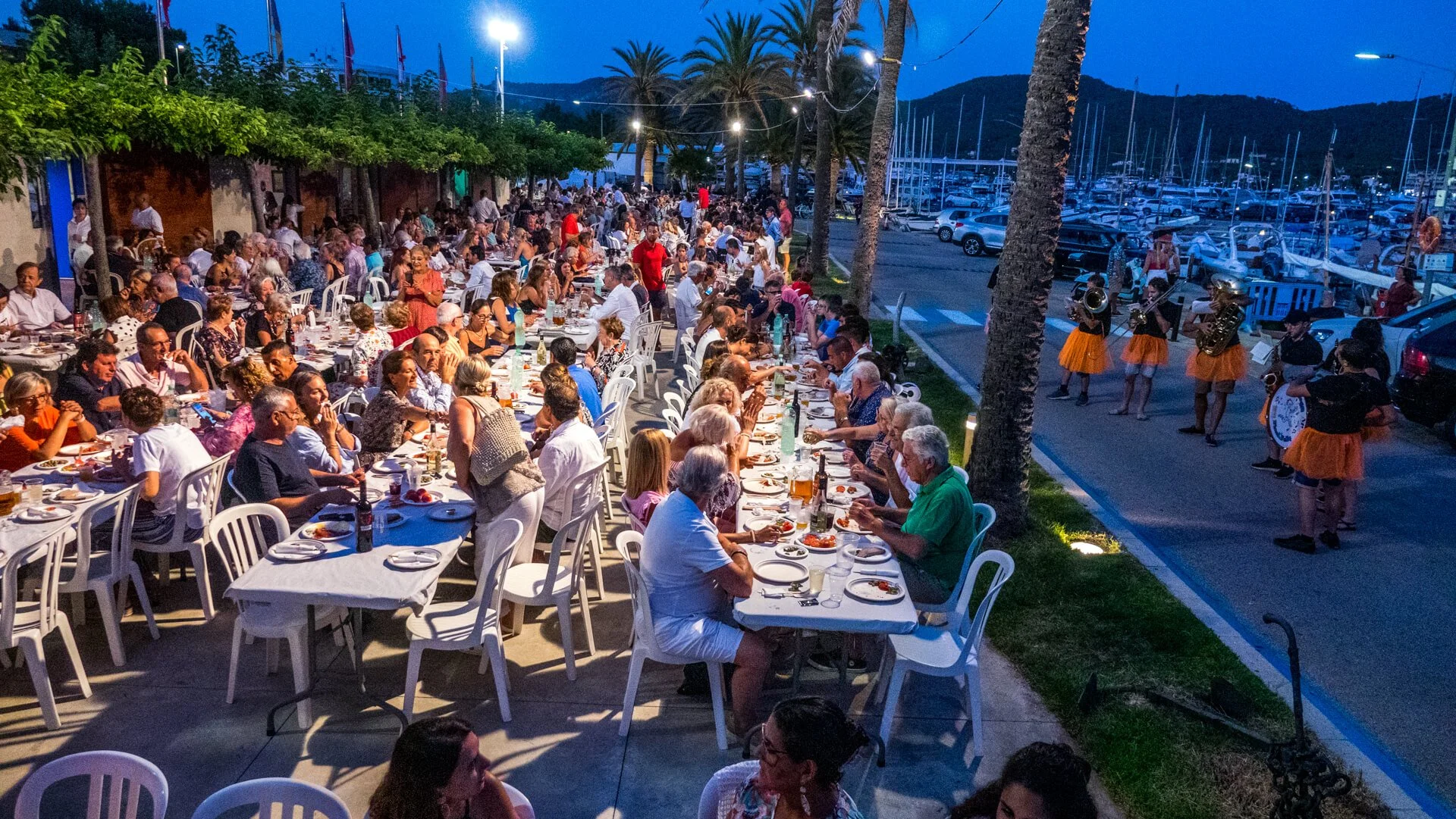 Una gran reunión al aire libre en la noche con muchas mesas llenas de personas comiendo y bebiendo, decoraciones con luces, palmeras y un mar con barcos en el fondo.