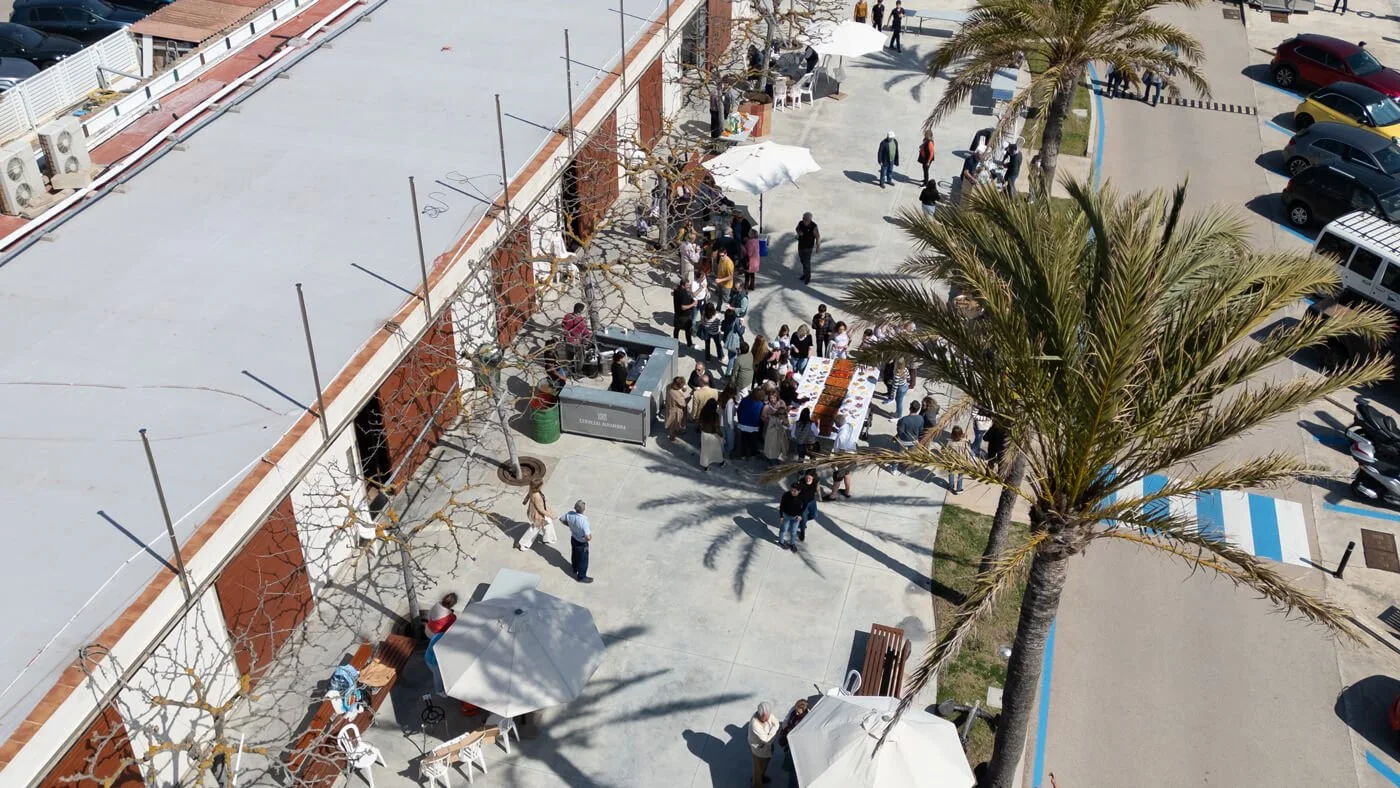 Reunión de personas en un evento al aire libre con una mesa de comida en una plaza con palmeras y árboles, estacionamiento en el fondo y sombra de sombrillas.