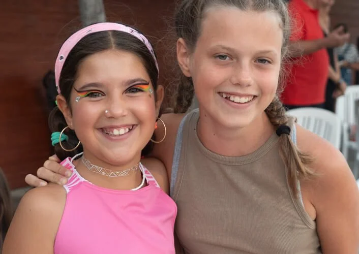 Dos niñas sonriendo y abrazándose en un ambiente interior, una lleva camiseta rosa y maquillaje colorido, la otra lleva camiseta beige y tiene el cabello en ponytail.