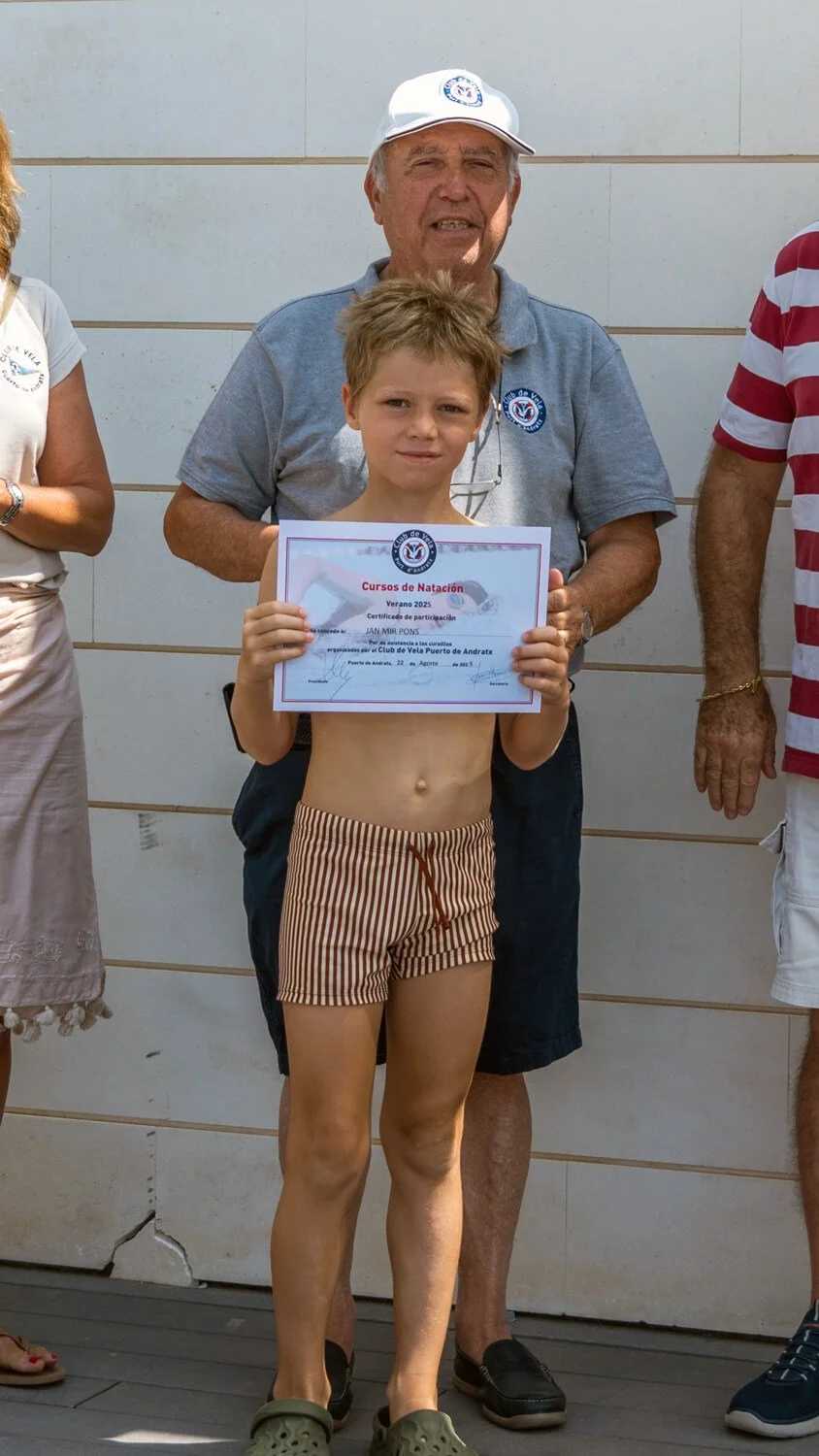 Un niño de pie con poca ropa y sandalias sostiene un certificado de participación en un curso de natación, acompañado de un adulto mayor con camiseta gris, sombrero y gafas, frente a una pared de color claro.