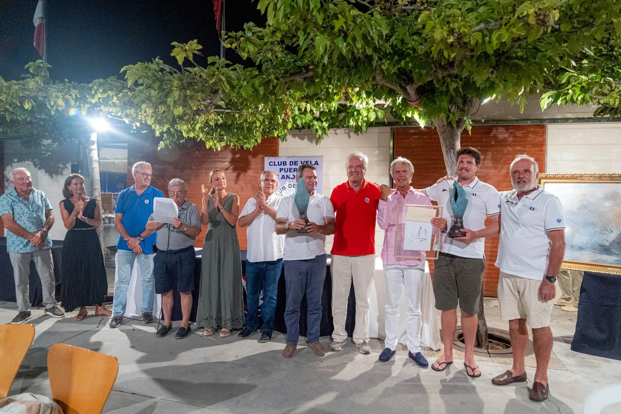 Un grupo de personas en una ceremonia al aire libre, algunas con premios y certificados, celebrando por la noche junto a un árbol y banderas de fondo.