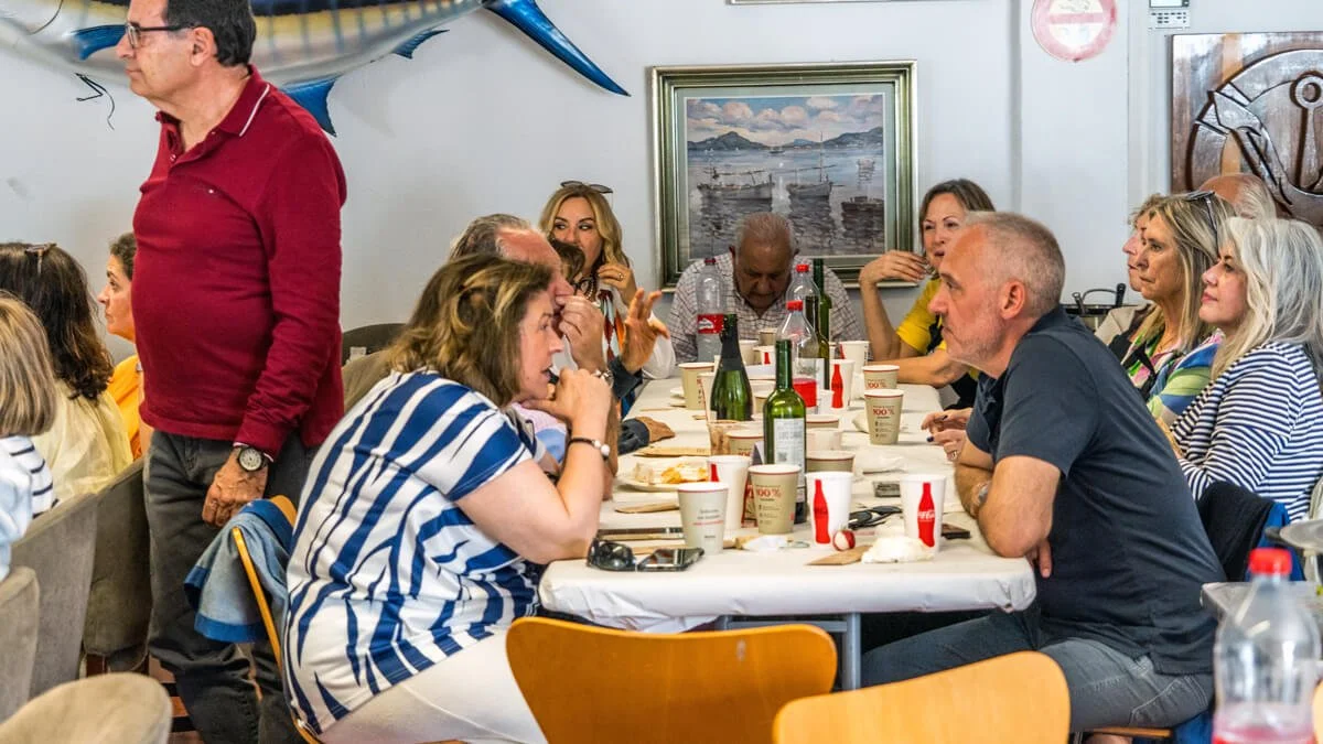 Grupo de personas sentadas en una mesa en un restaurante, conversando. Hay botellas, vasos, platos y una manta en la mesa, y un cuadro y una escultura en la pared de fondo.