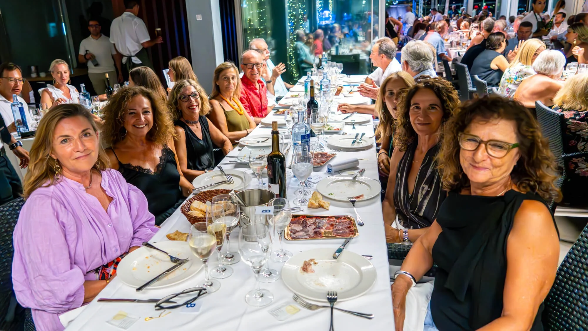 Un grupo de personas celebrando en una comida en un restaurante con muchas mesas llenas de comida, con una ventana grande y decoraciones de luz en el fondo.