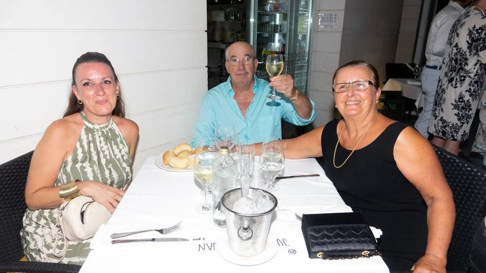 Tres personas, dos mujeres y un hombre, sentadas en una mesa en un restaurante, brindando con vino. La mesa tiene comida, copas y un cubo con hielo.
