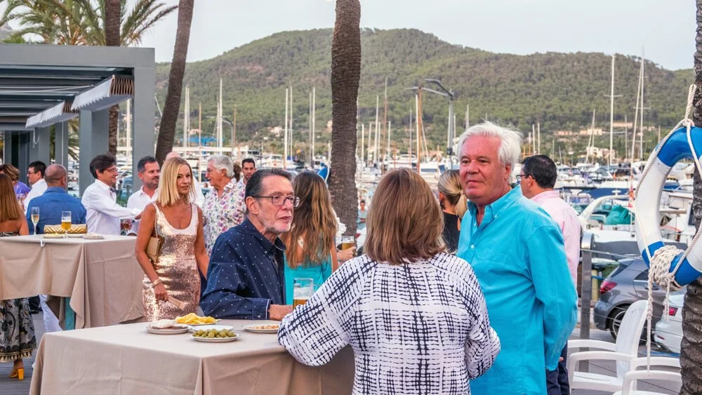 Personas socializando en un evento al aire libre cerca del puerto con barcos y montañas en el fondo.