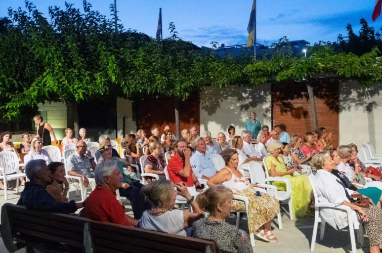 Muchas personas adultas sentadas en sillas en un evento al aire libre en la noche, con árboles y banderas en el fondo.