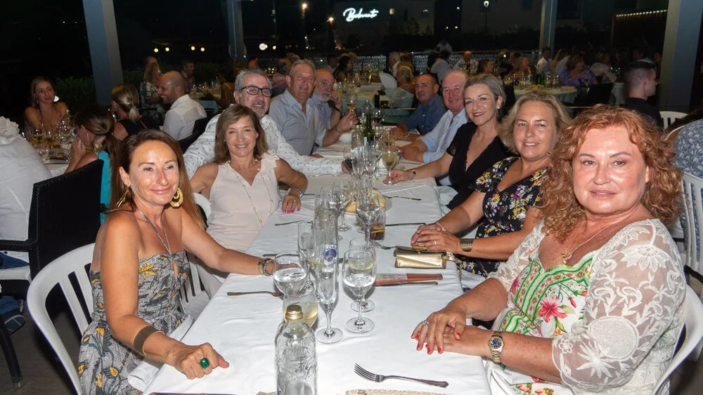 Grupo de personas sentadas en una mesa durante una cena en un restaurante, sonriendo y disfrutando de la compañía, fondo con más mesas y decoración luminosa.
