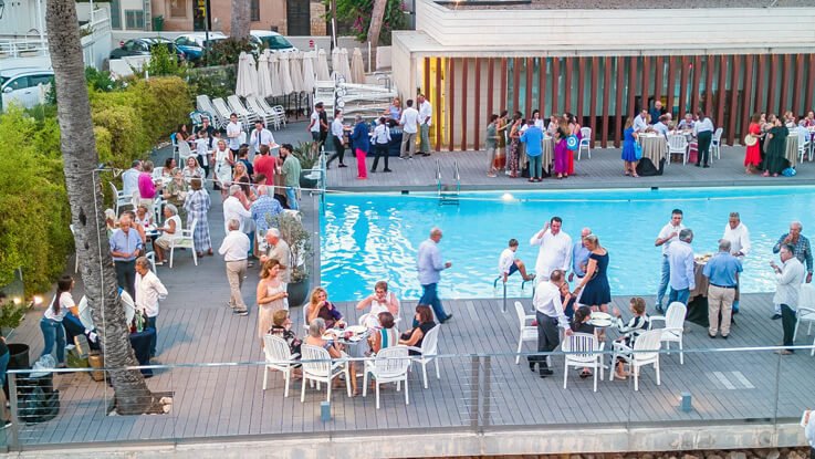 Reunión social en una piscina con muchas personas, algunas en la terraza y otras en las mesas, en un ambiente al aire libre con vegetación y estructuras modernas.