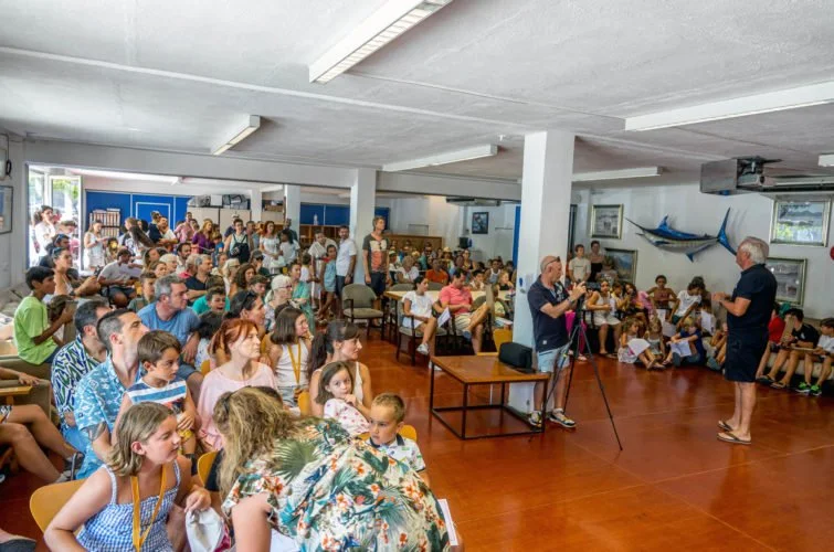 Una sala llena de niños y adultos en una presentación o taller, con dos hombres en el frente, uno con una cámara y otro con un micrófono, en un ambiente decorado con un pez grande en la pared.