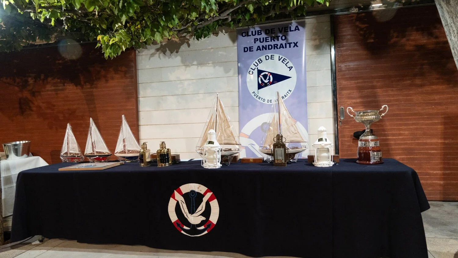 Mesa decorada con modelos de barcos a vela, velas y trofeos, bajo una pancarta del Club de Vela Puerto de Andraitx, en un evento náutico.