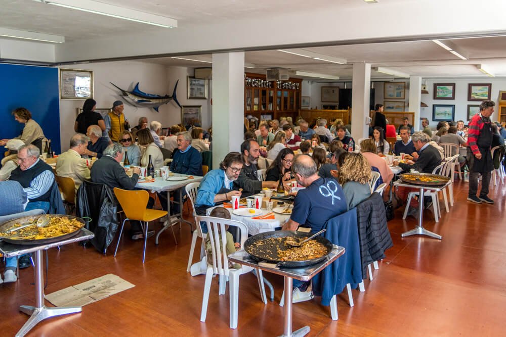 Personas en un restaurante comiendo, con algunos platillos grandes de comida española como paella en las mesas y paredes decoradas con pinturas y un modelo de tiburón en la pared.