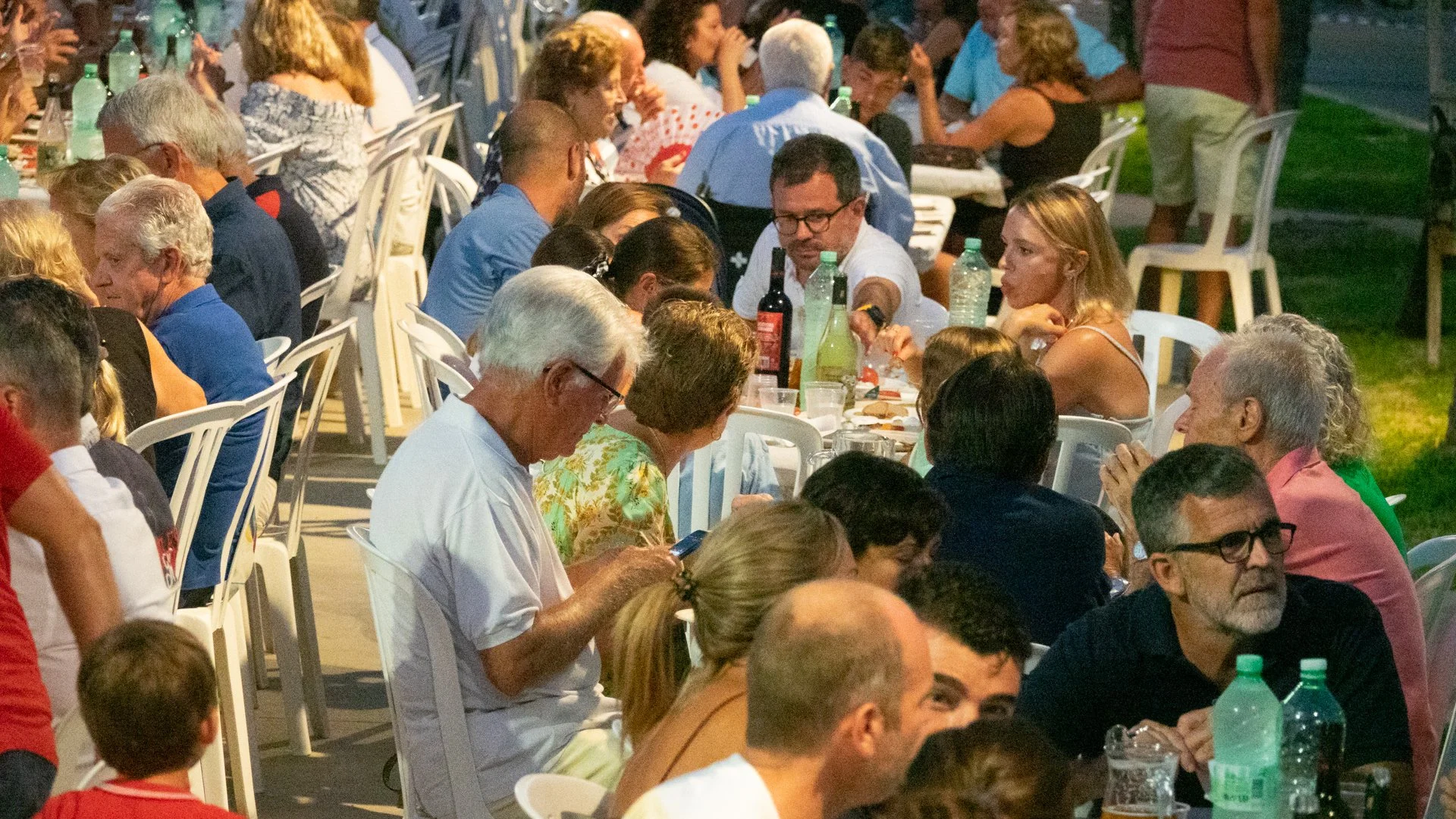Grupo de personas sentadas en una mesa al aire libre en una reunión social, con bebidas y comida, en un ambiente nocturno.