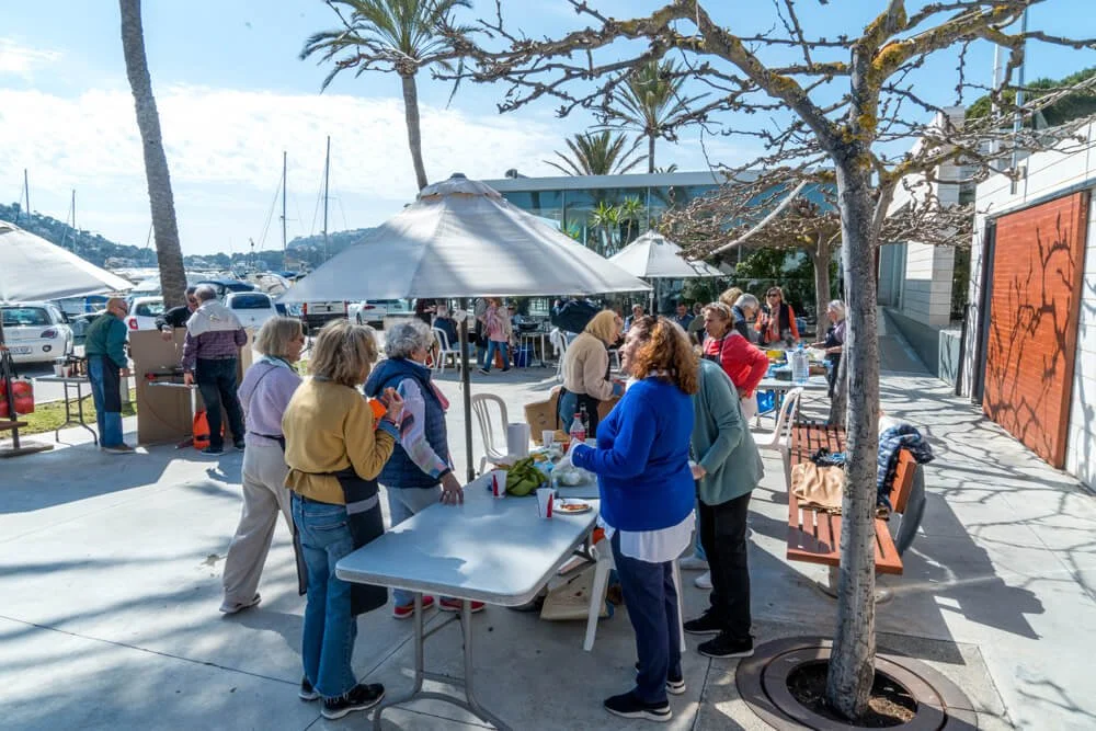 Grupo de personas mayores disfrutando de una comida o reunión al aire libre en un área con mesas, sombrillas y árboles en un muelle o puerto.