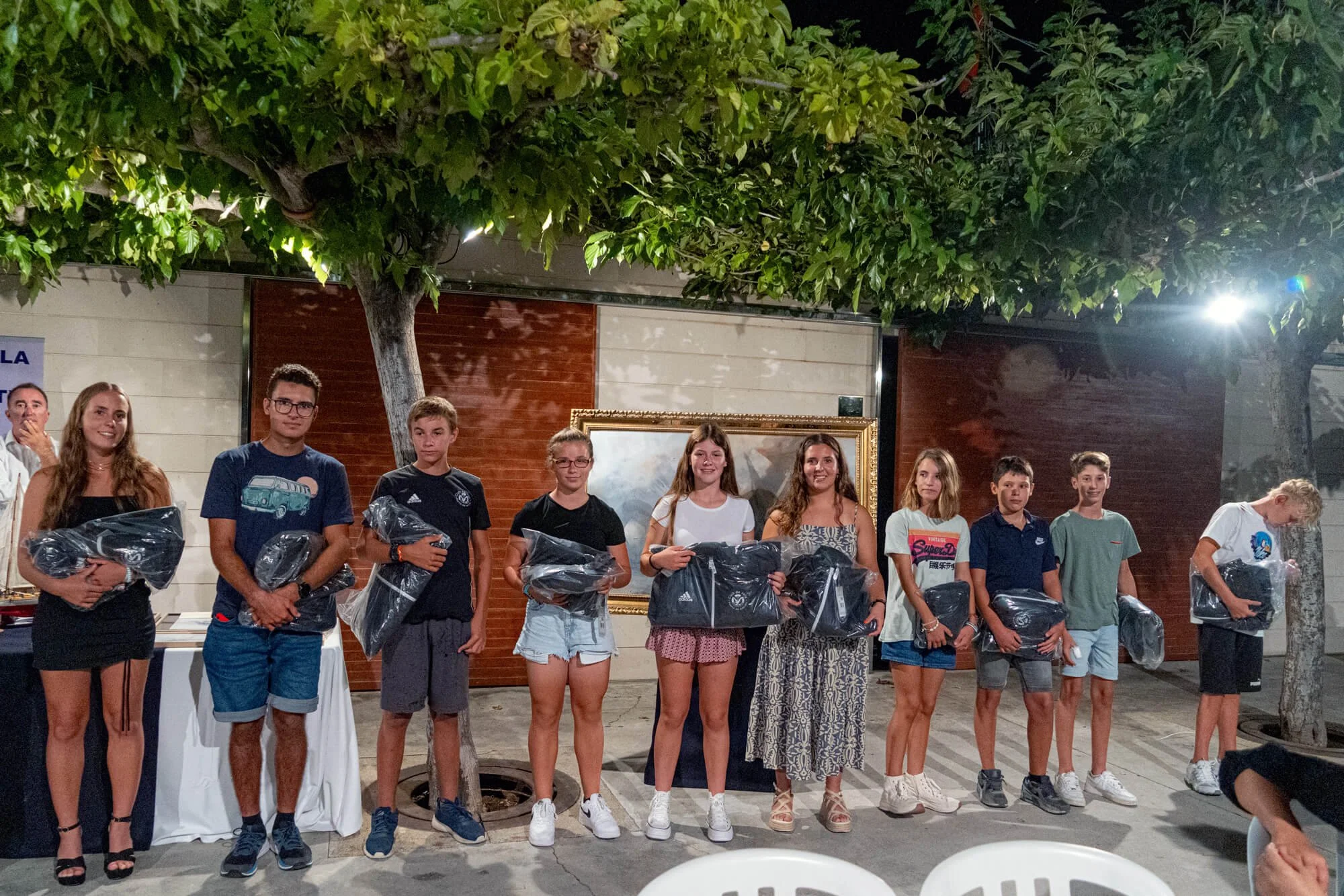 Un grupo de niños y jóvenes posando con premios en una ceremonia en un espacio al aire libre, con árboles y luces.