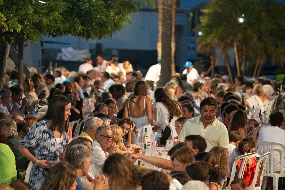 Un gran grupo de personas en una comida al aire libre, sirviéndose y disfrutando de la cena en una reunión familiar o social durante la noche.