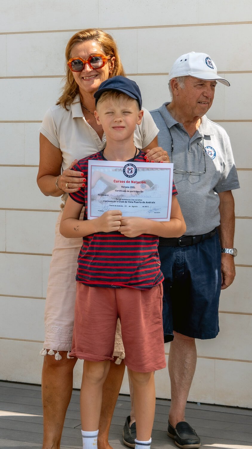 Un niño sostiene un certificado de participación en cursos de natación, acompañado por una mujer y un hombre, en un evento al aire libre.