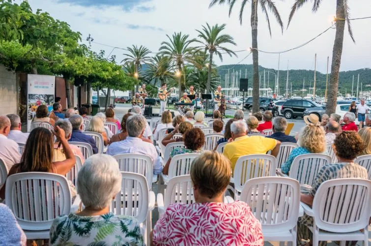 Concierto al aire libre en la costa, con público sentado en sillas blancas y músicos tocando en un escenario con palmeras y yates en el fondo durante el atardecer.