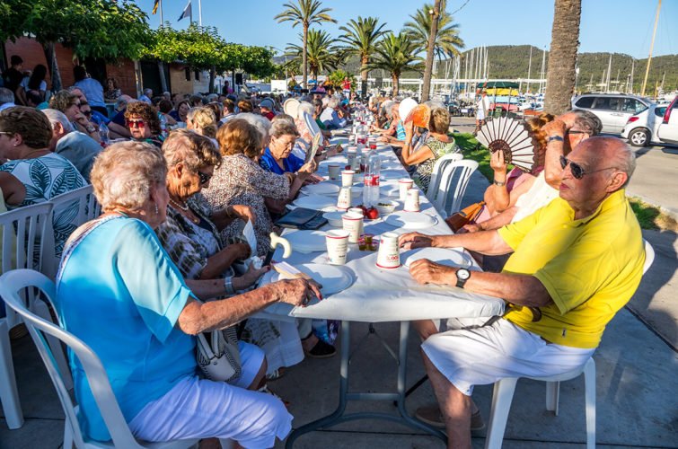 Grupo de personas mayores sentadas en una mesa larga al aire libre durante una reunión social, con palmeras y barcos en el fondo.