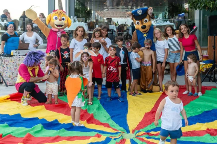 Niños y adultos en una fiesta infantil en un área con un paracaídas colorido, acompañados de personajes disfrazados de dibujos animados. Algunos niños están siendo atendidos por un payaso con peluca púrpura.