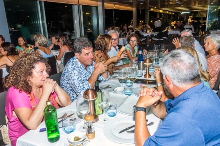 Personas sentadas en una mesa de restaurante, algunas leyendo, otras conversando, con bebidas y comida en un ambiente elegante y bien iluminado.