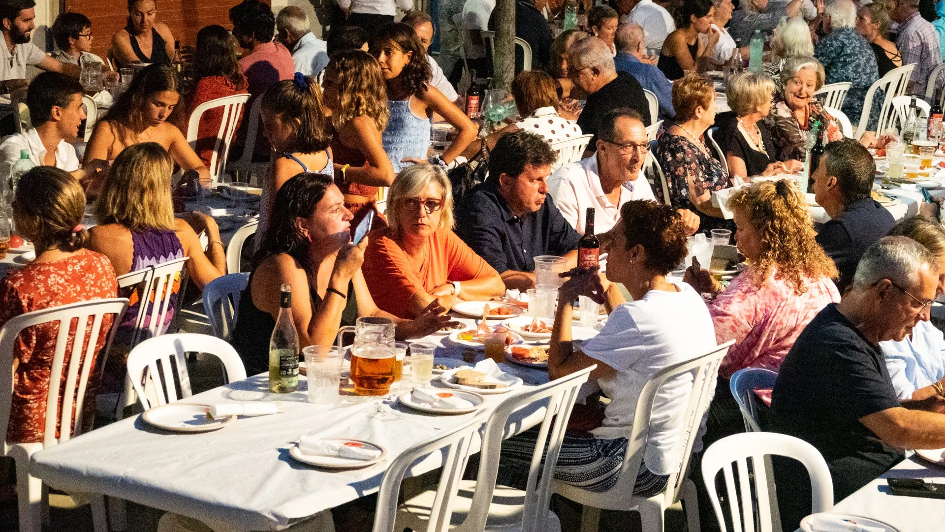 Grupo de personas sentadas alrededor de mesas en una reunión o evento social al aire libre por la noche.