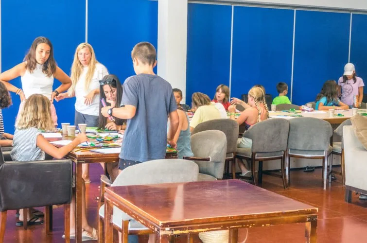 Niños y adolescentes en una sala con mesas y sillas, haciendo manualidades y participando en actividades grupales.