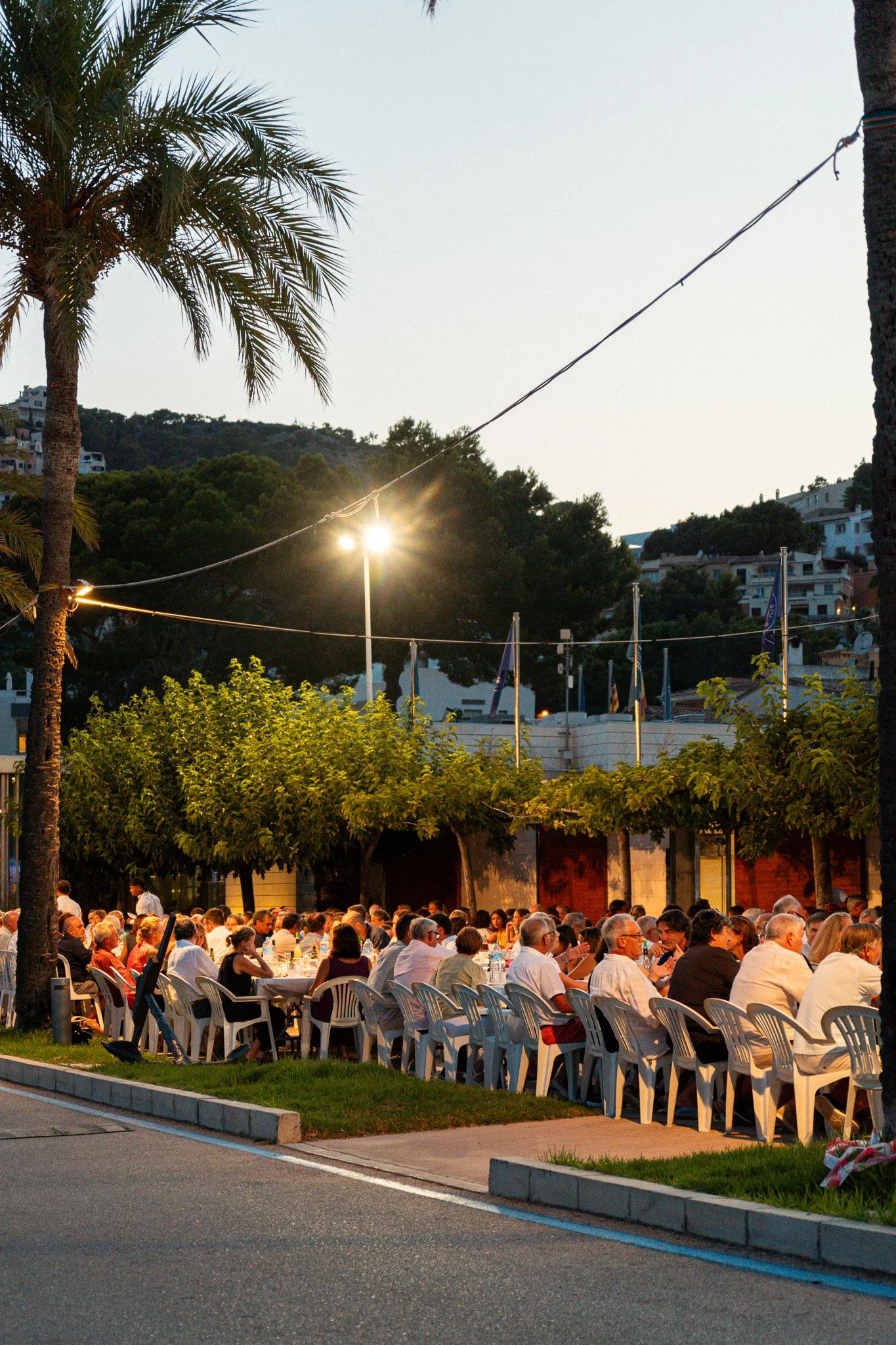 Reunión al aire libre durante el atardecer con muchas personas sentadas en mesas, árboles y palmeras rodean el lugar, y en el fondo colinas con casas.