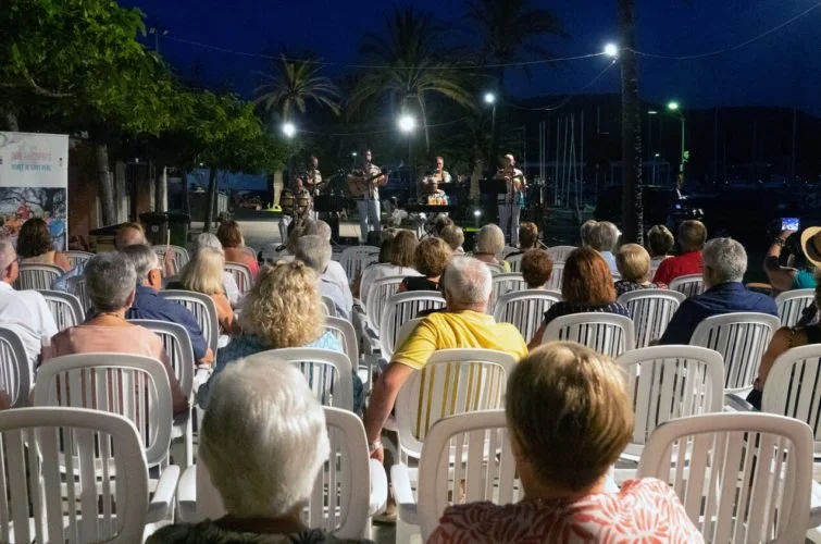 Concierto al aire libre con banda musical en el escenario y público sentado en sillas blancas en la noche, rodeada de palmeras.