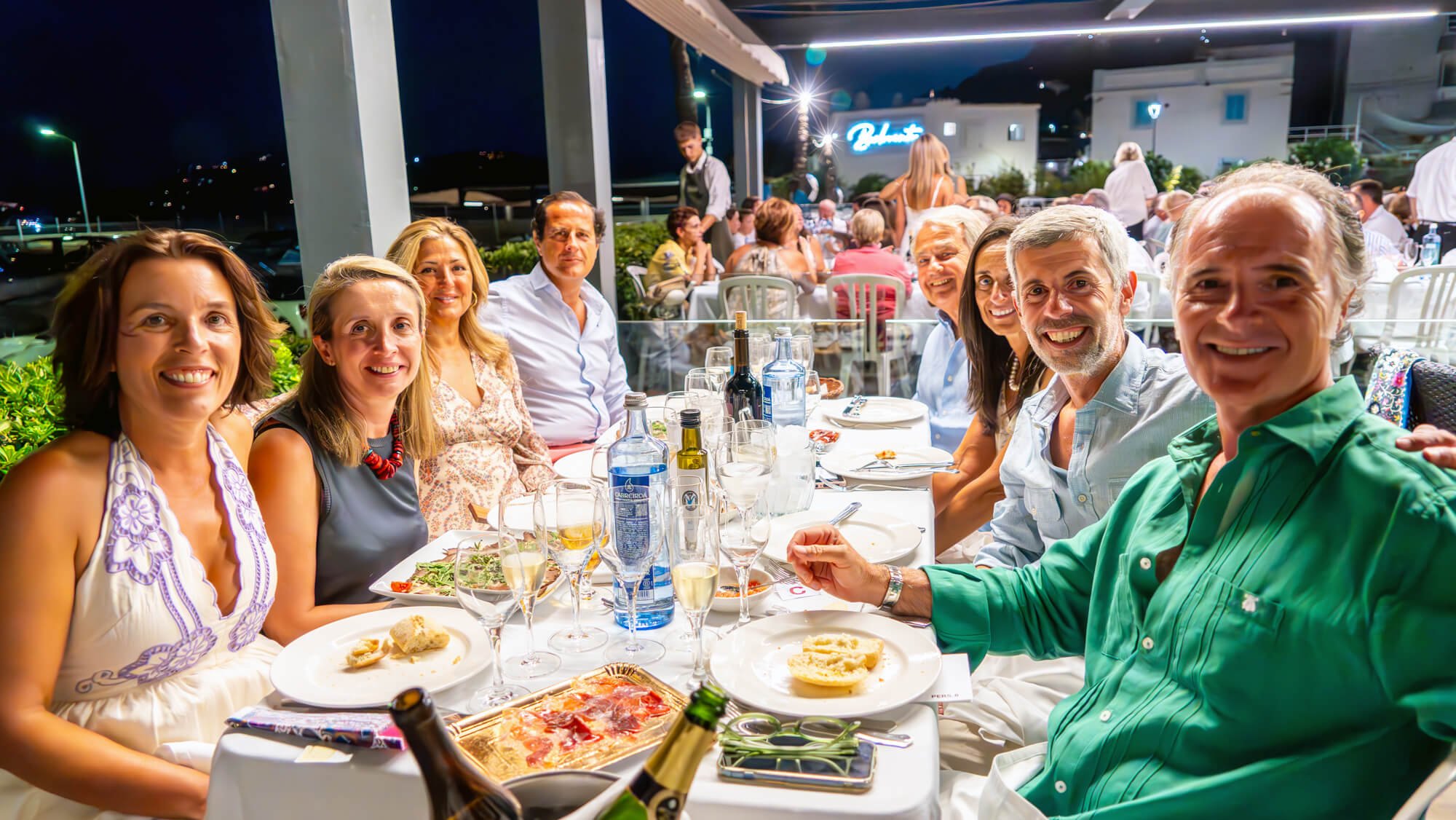 Grupo de personas sentadas en una mesa de cena al aire libre en la noche, con platos, copas, botellas de vino y cerveza, en un restaurante con ambiente nocturno y edificios iluminados en el fondo.