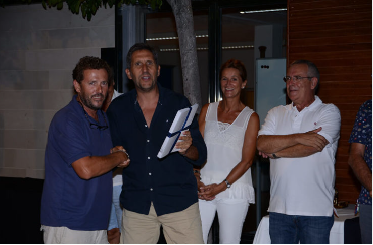 Cuatro personas en una reunión al aire libre, entre ellas un hombre con camisa azul sosteniendo un documento, y otra mujer con blusa blanca sonriendo, mientras un hombre con camiseta blanca con los brazos cruzados los observa.