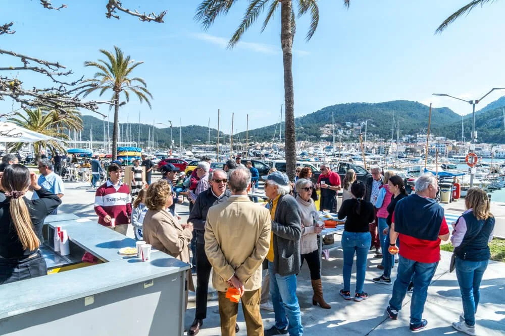 Grupo de personas en un muelle con palmeras, barcos y montañas al fondo, disfrutando de una actividad social al aire libre en un día soleado.