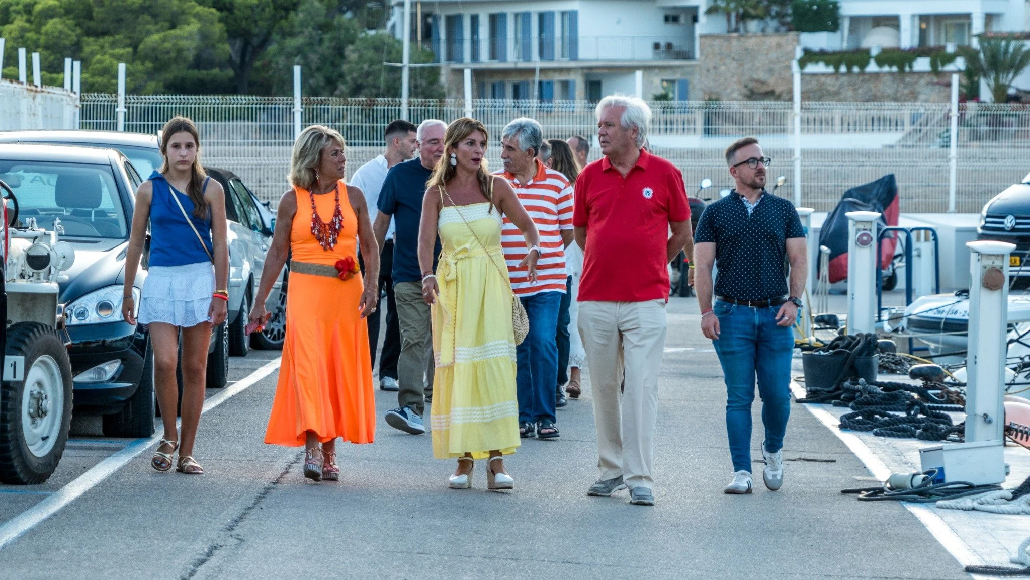 Grupo de personas caminando en un estacionamiento con autos y árboles al fondo, vestidas con ropa casual y elegante.