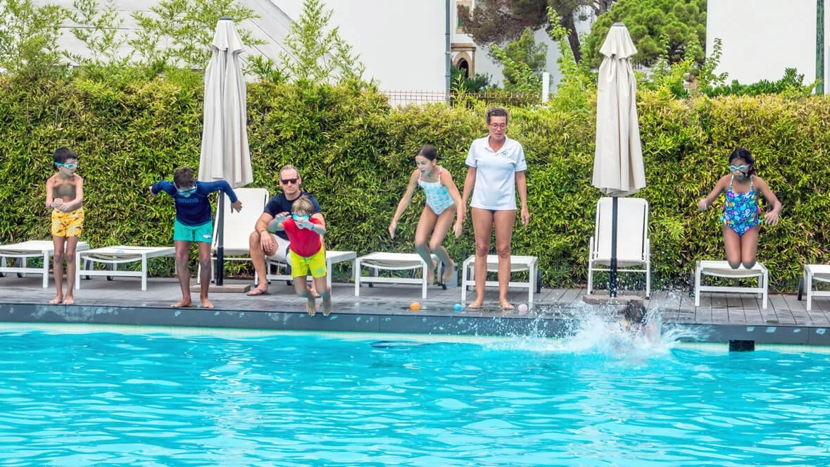 Niños y adultos en una alberca, algunos niños saltando dentro del agua y otros en las bancas junto a la piscina, con vegetación de fondo.