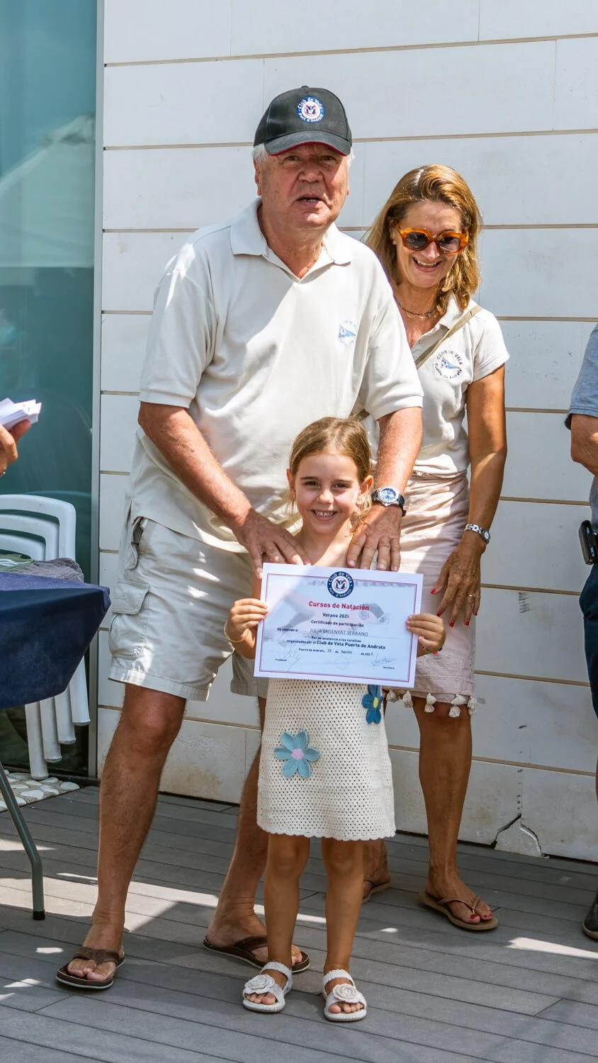 Una niña sostiene un certificado de participación en un curso de natación, mientras es felicitada por un hombre mayor vestido con camiseta beige y gorra, y una mujer con gafas de sol y blusa blanca, en un evento al aire libre.
