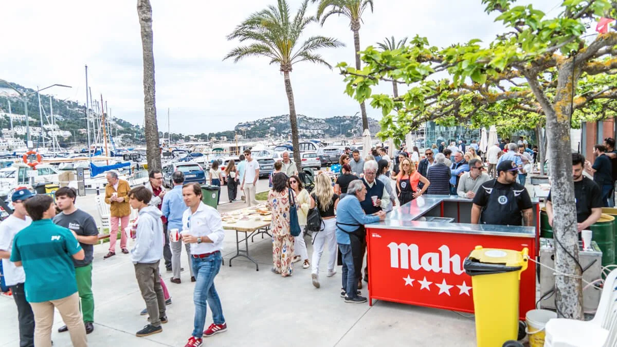 Personas en una reunión social al aire libre en un muelle junto a yates, con mesas y un puesto de comida con el cartel 'Mah'.