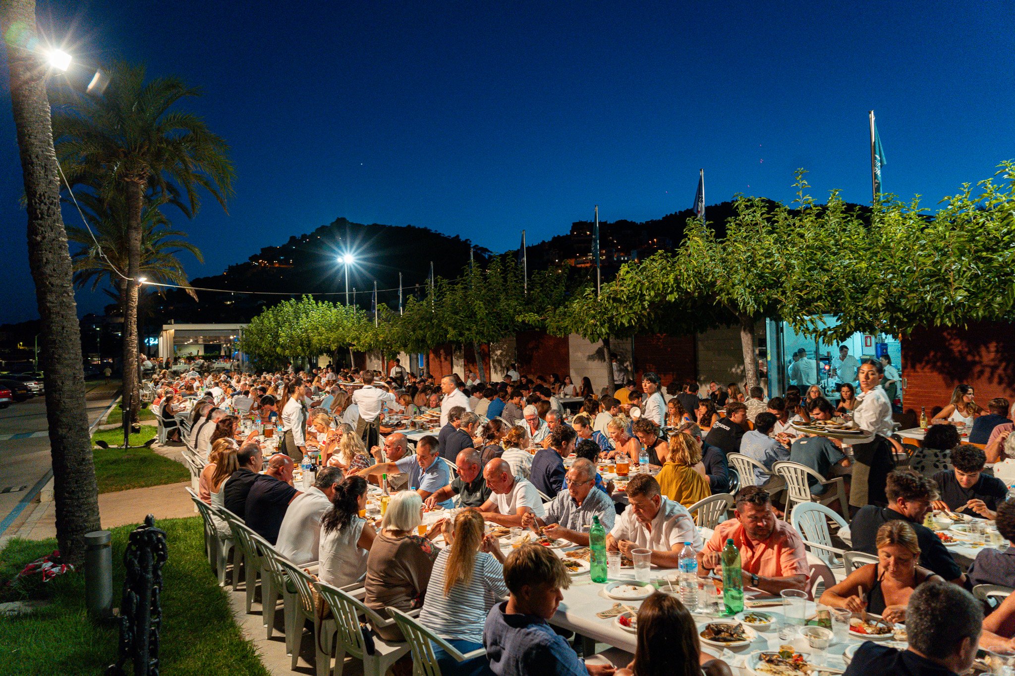 Una gran reunión al aire libre durante la noche con muchas mesas y personas comiendo, con palmeras y colinas en el fondo y luces en las colinas.