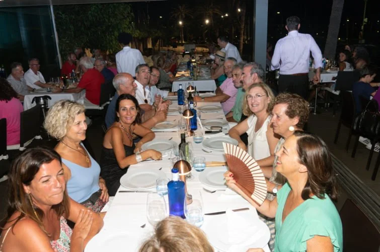 Grupo de personas sentadas en una mesa larga en un restaurante durante la noche, algunos conversando y otros sonriendo.