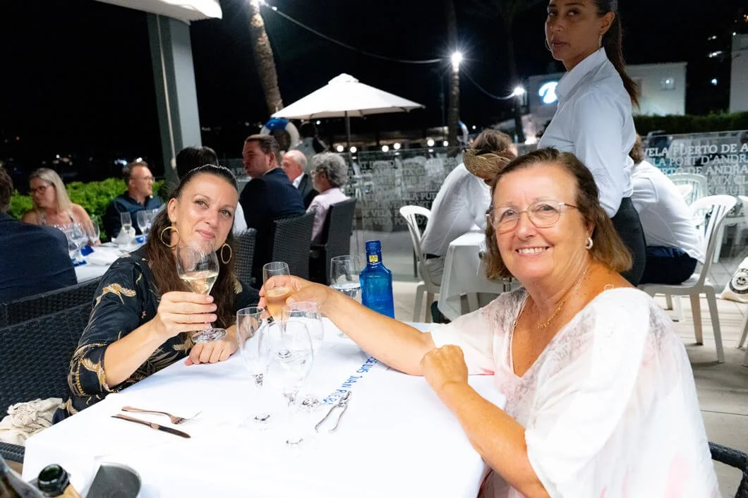 Dos mujeres adultas en una mesa de restaurante brindando con copas de vino, en un ambiente nocturno y elegante, con otros comensales y una camarera en el fondo.