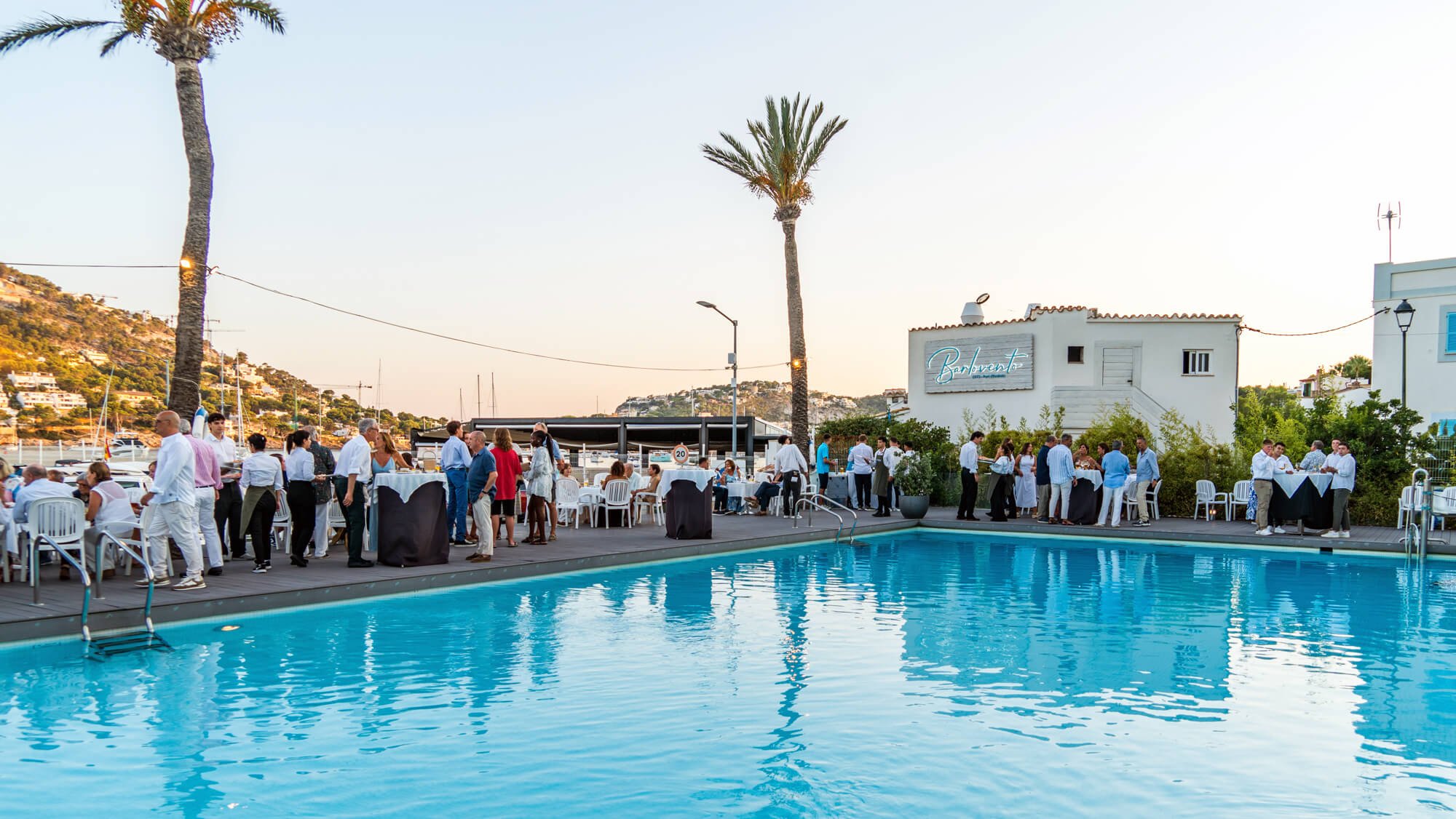 Un grupo de personas en una fiesta al aire libre junto a una piscina, con mesas y sillas, mientras el sol se pone y un paisaje de montañas con casas y barcos en el fondo.