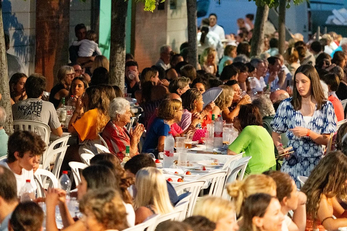 Una gran reunión de personas sentadas en una larga mesa al aire libre, comiendo y socializando, con algunos niños entre los adultos, en un ambiente diurno con árboles y edificios en el fondo.